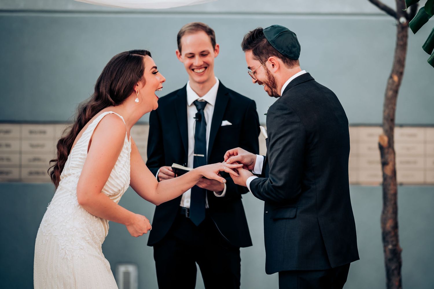 Couple exchanging rings during outdoor wedding ceremony with officiant under modern architectural backdrop.