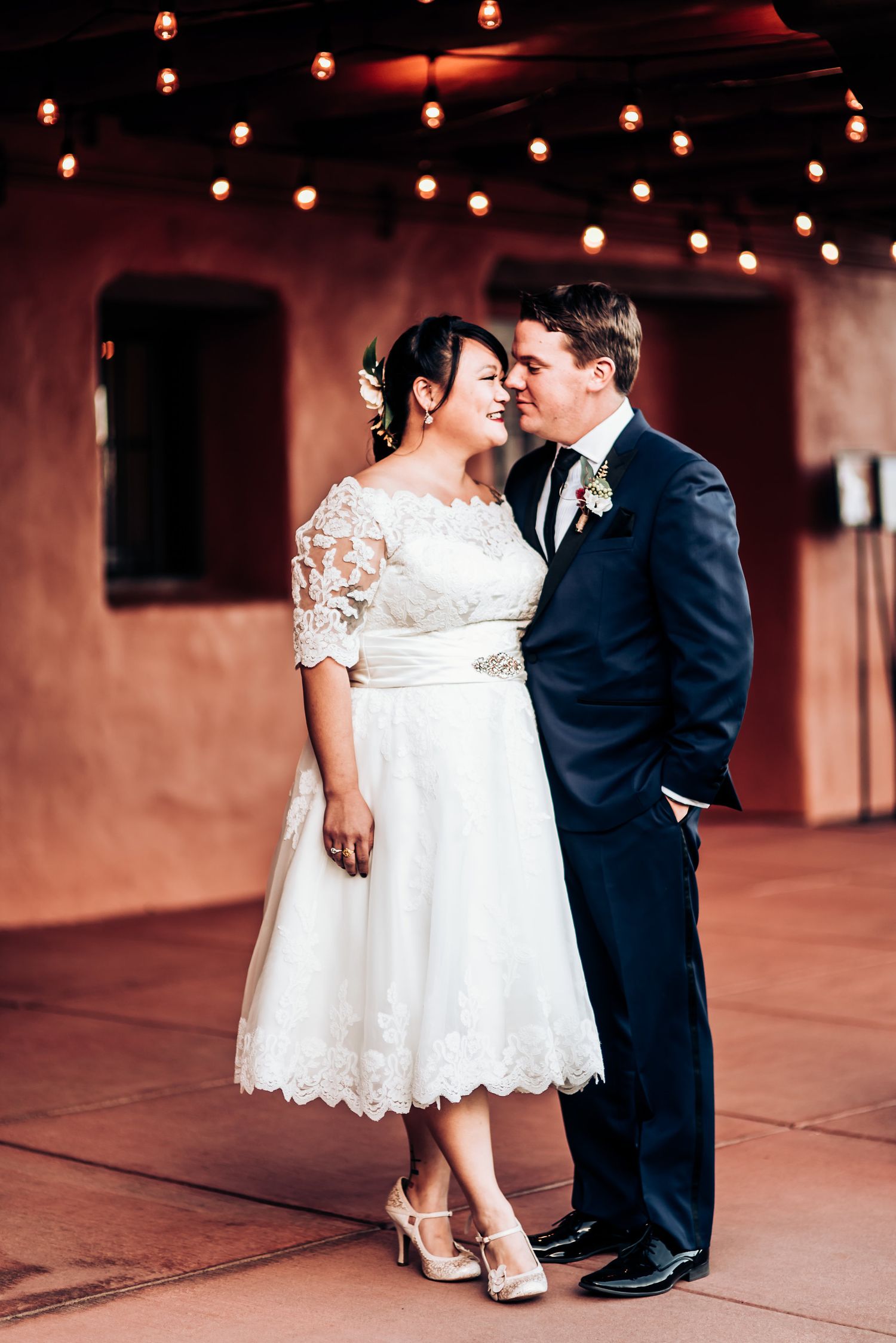 A romantic wedding couple pose under string lights on a terrace, the bride wearing a tea-length lace dress.