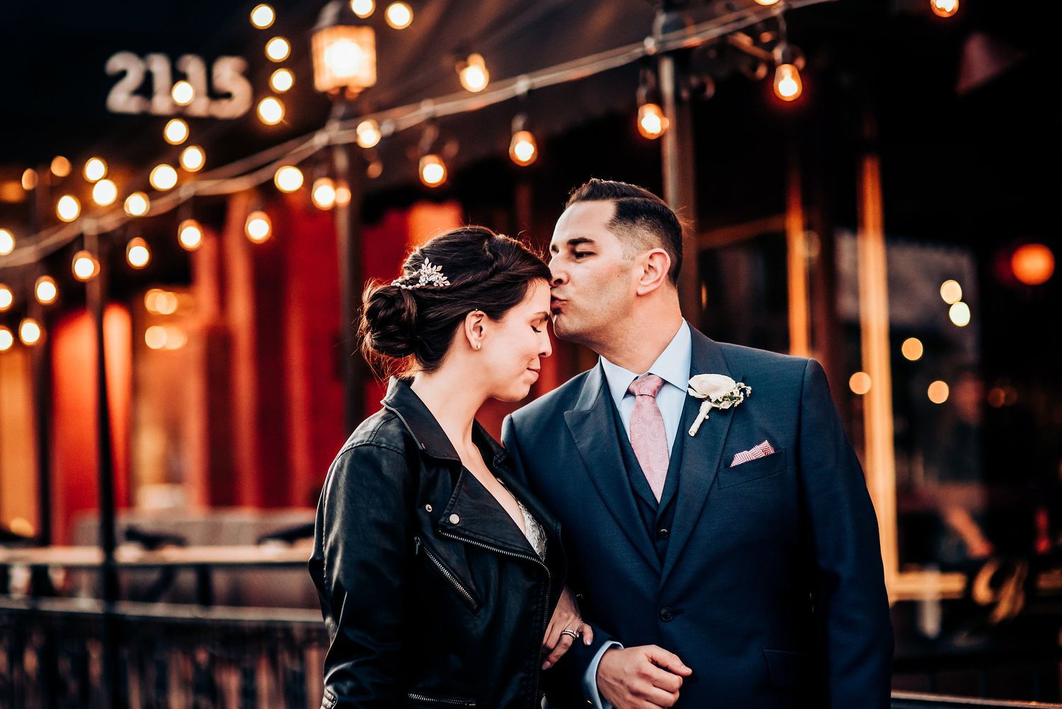 A romantic couple shares an intimate moment under glowing string lights outside a restaurant.