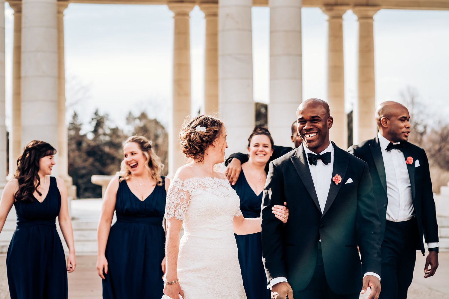 Wedding party in navy and white walking through an elegant colonnade during golden hour.