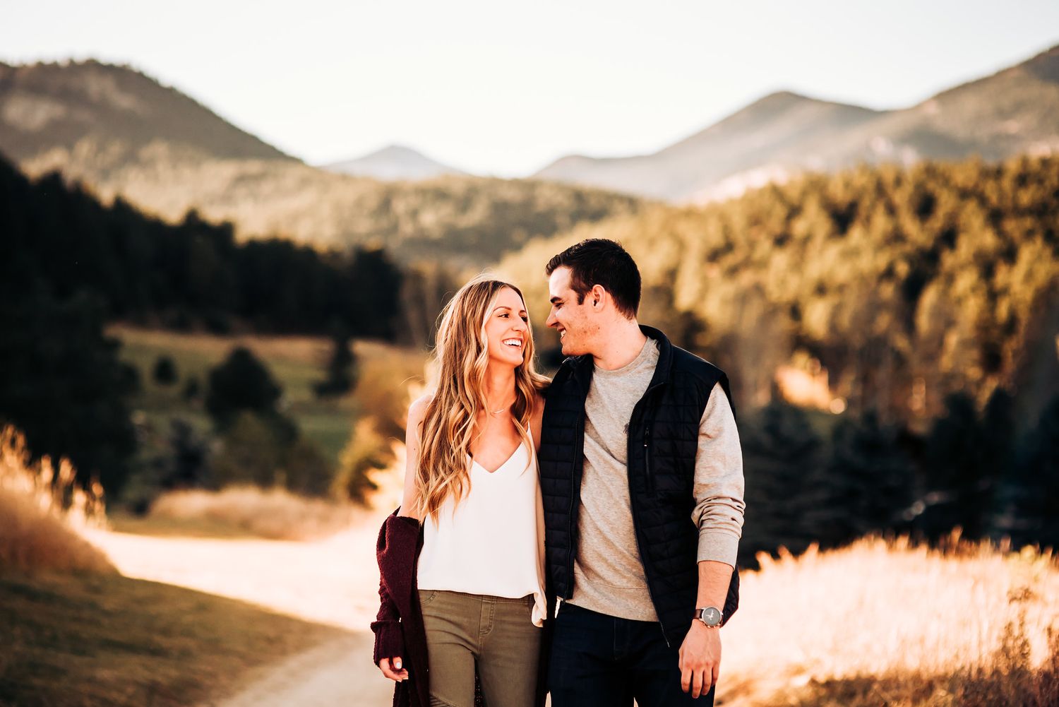 A couple shares an intimate moment on a mountain trail with rolling hills in the background.