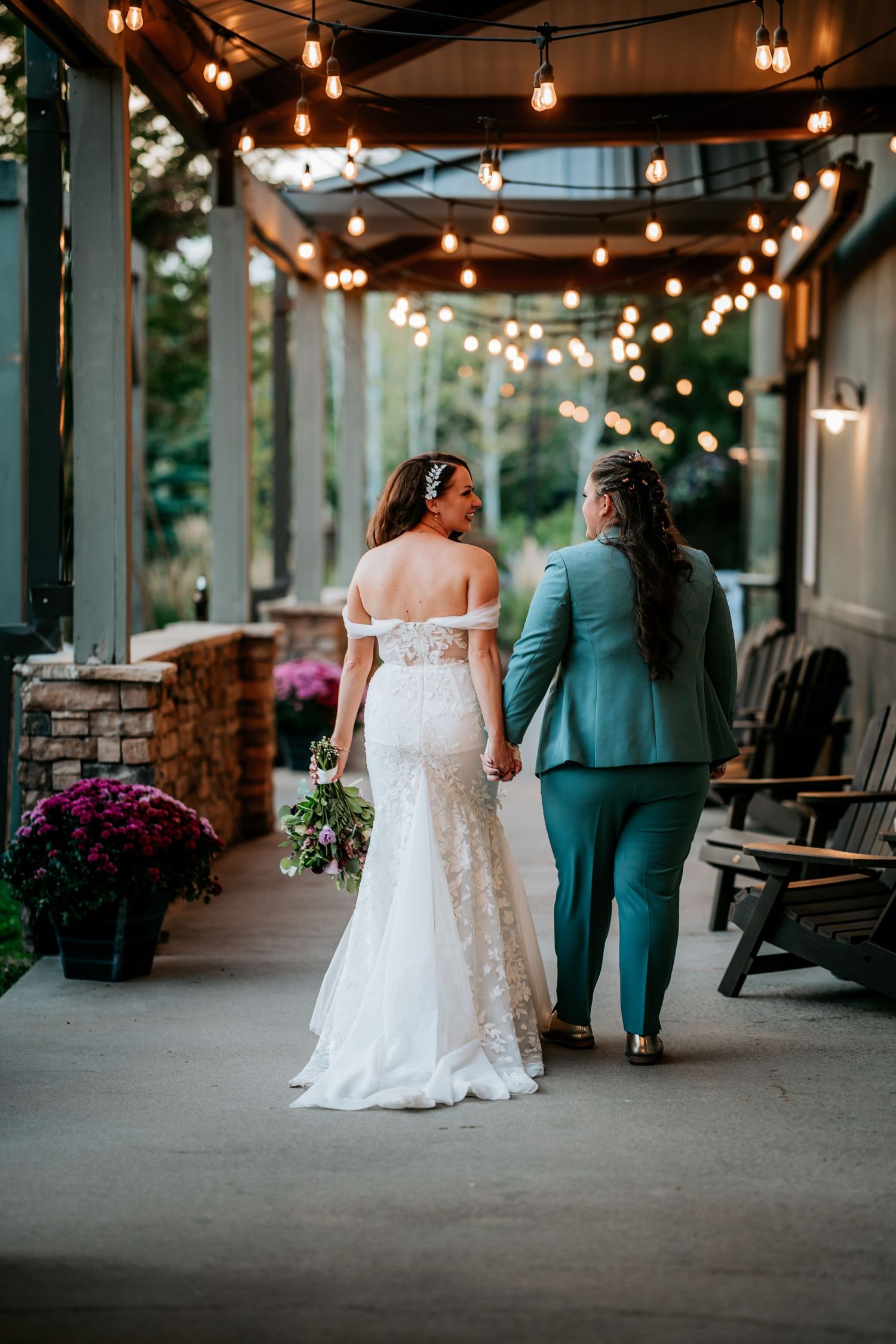 Two brides walk hand-in-hand under string lights on a patio, one in white lace and one in teal.