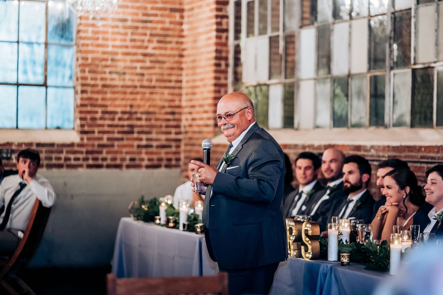 A speaker delivers a toast during wedding reception while guests listen from decorated tables.