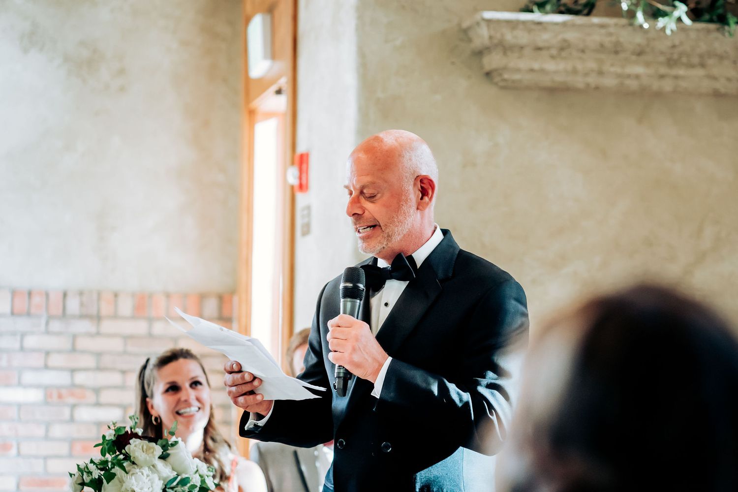 A speaker in a black tuxedo gives an emotional wedding toast while holding a microphone.