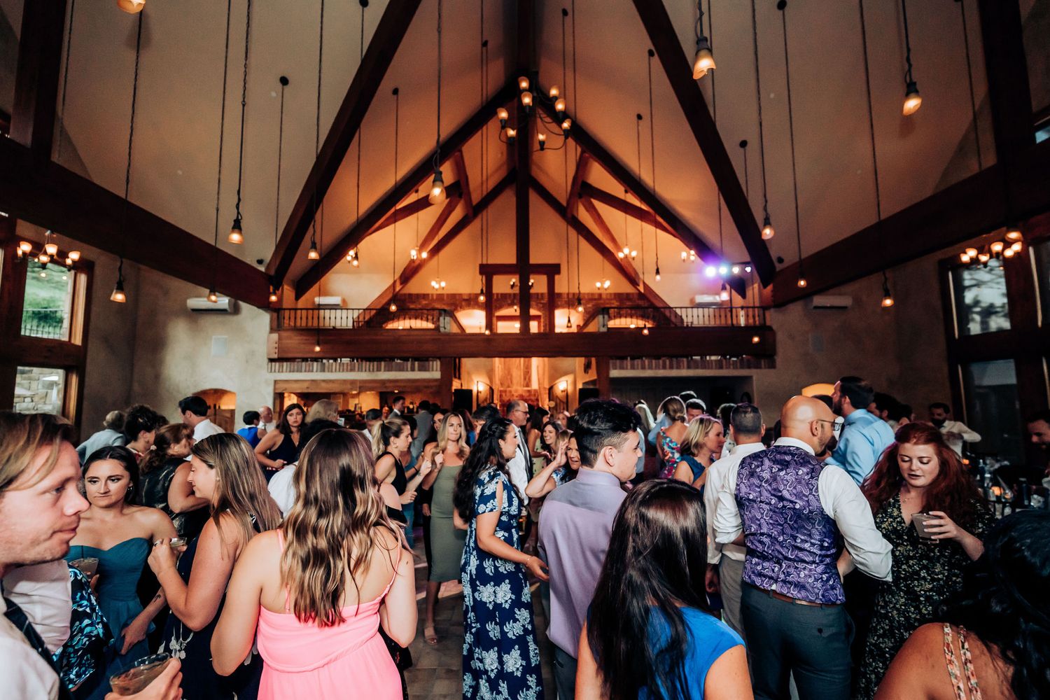 Large group of wedding guests dancing beneath vaulted wooden ceiling with hanging lights.