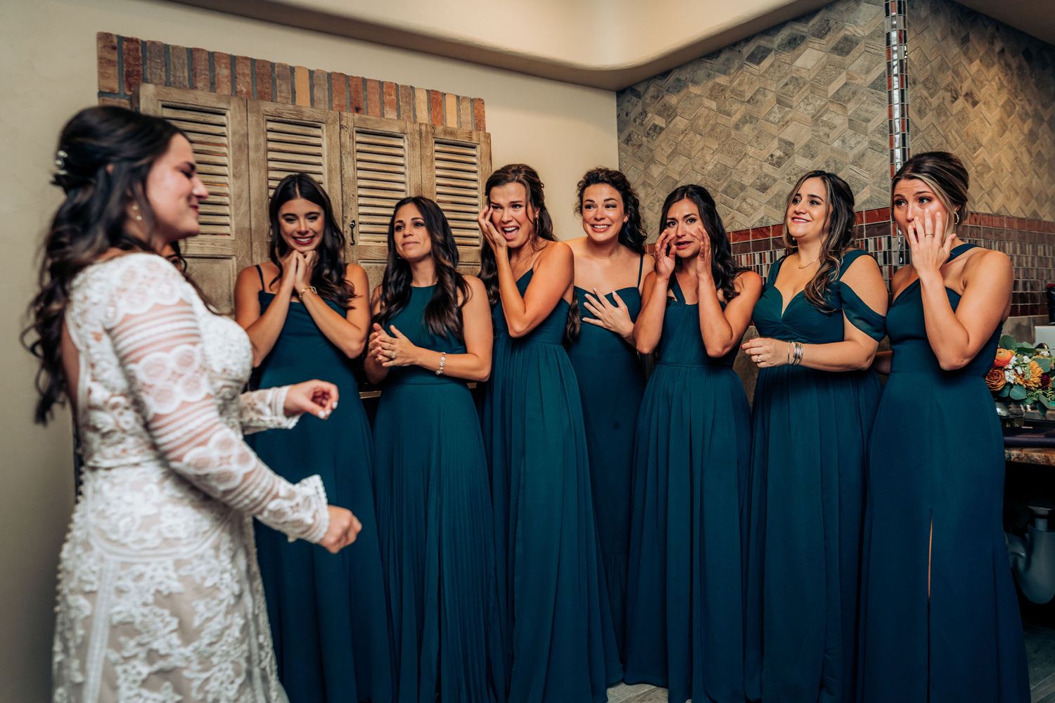 Bridesmaids in teal dresses share emotional reactions with bride in white lace gown.