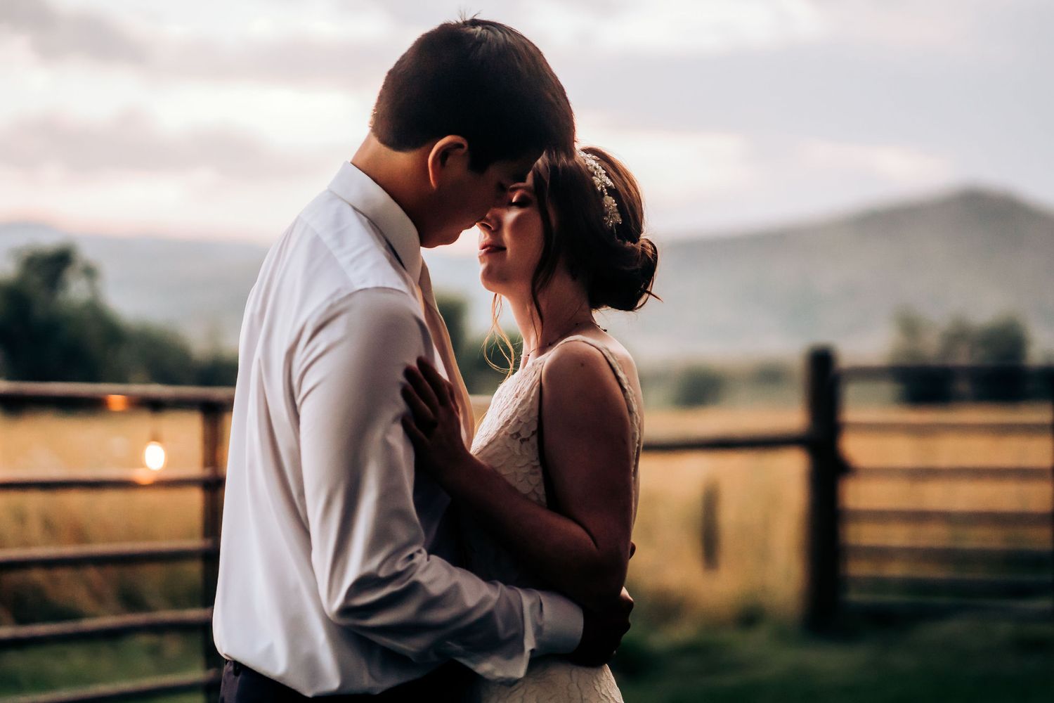 A romantic couple shares an intimate moment at sunset against a rustic countryside backdrop with wooden fencing.
