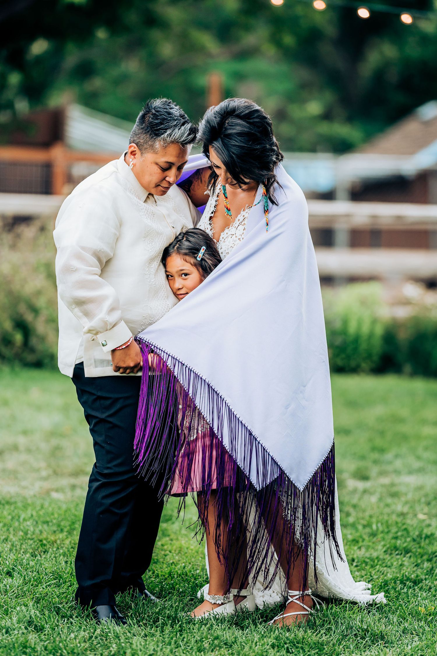 A tender family moment shared under a white shawl with fringes in an outdoor garden setting.