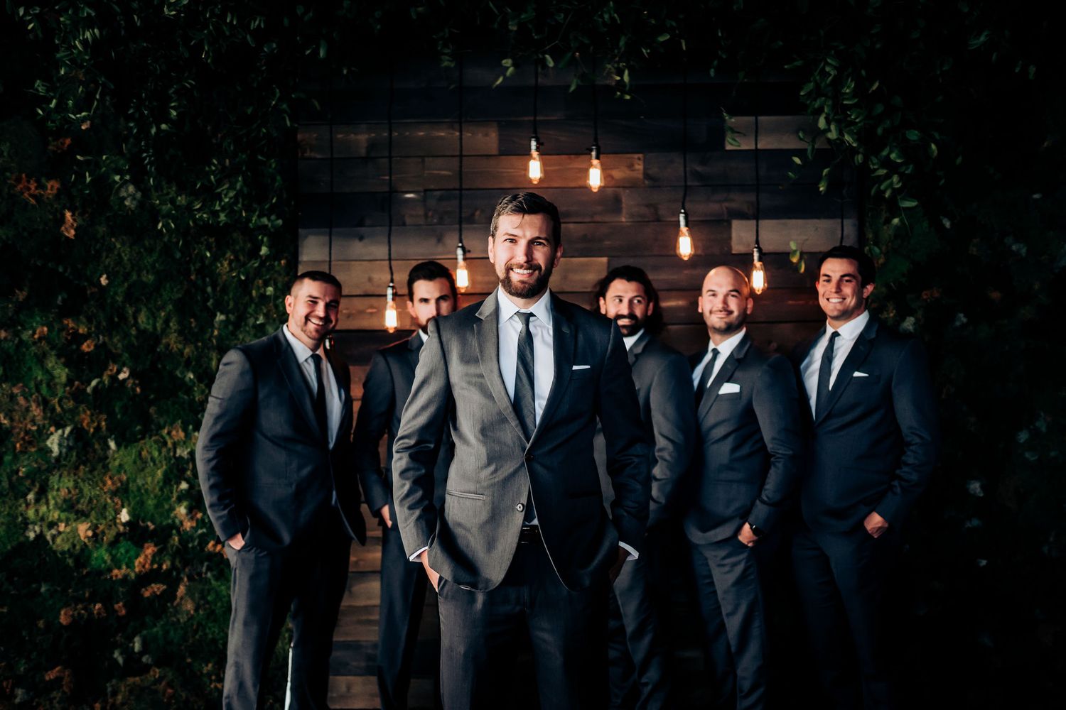 Groomsmen pose for a stylish portrait against a rustic brick wall with hanging lights and greenery.