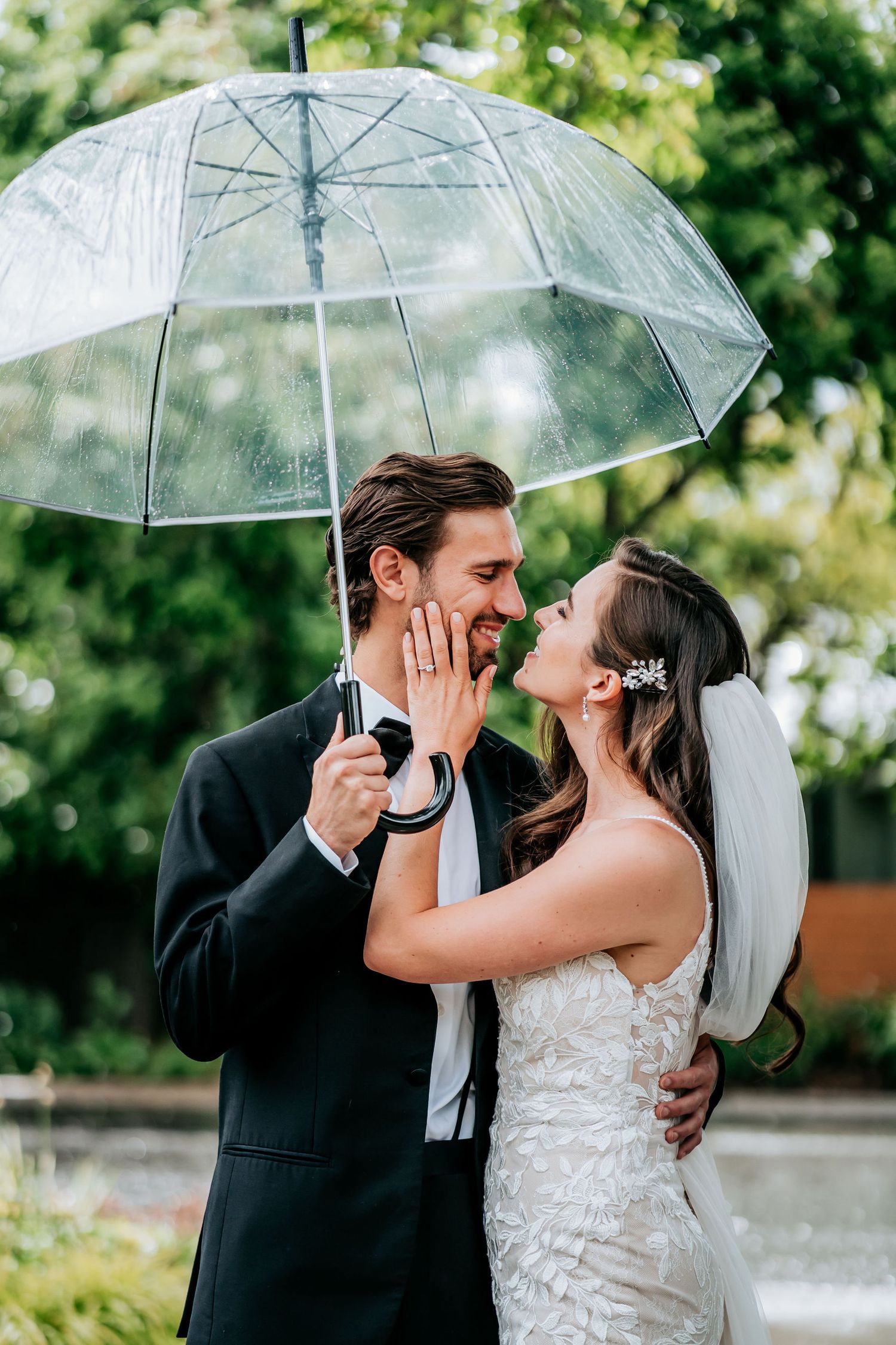 A romantic wedding couple shares a tender moment under a clear umbrella in a lush garden setting.