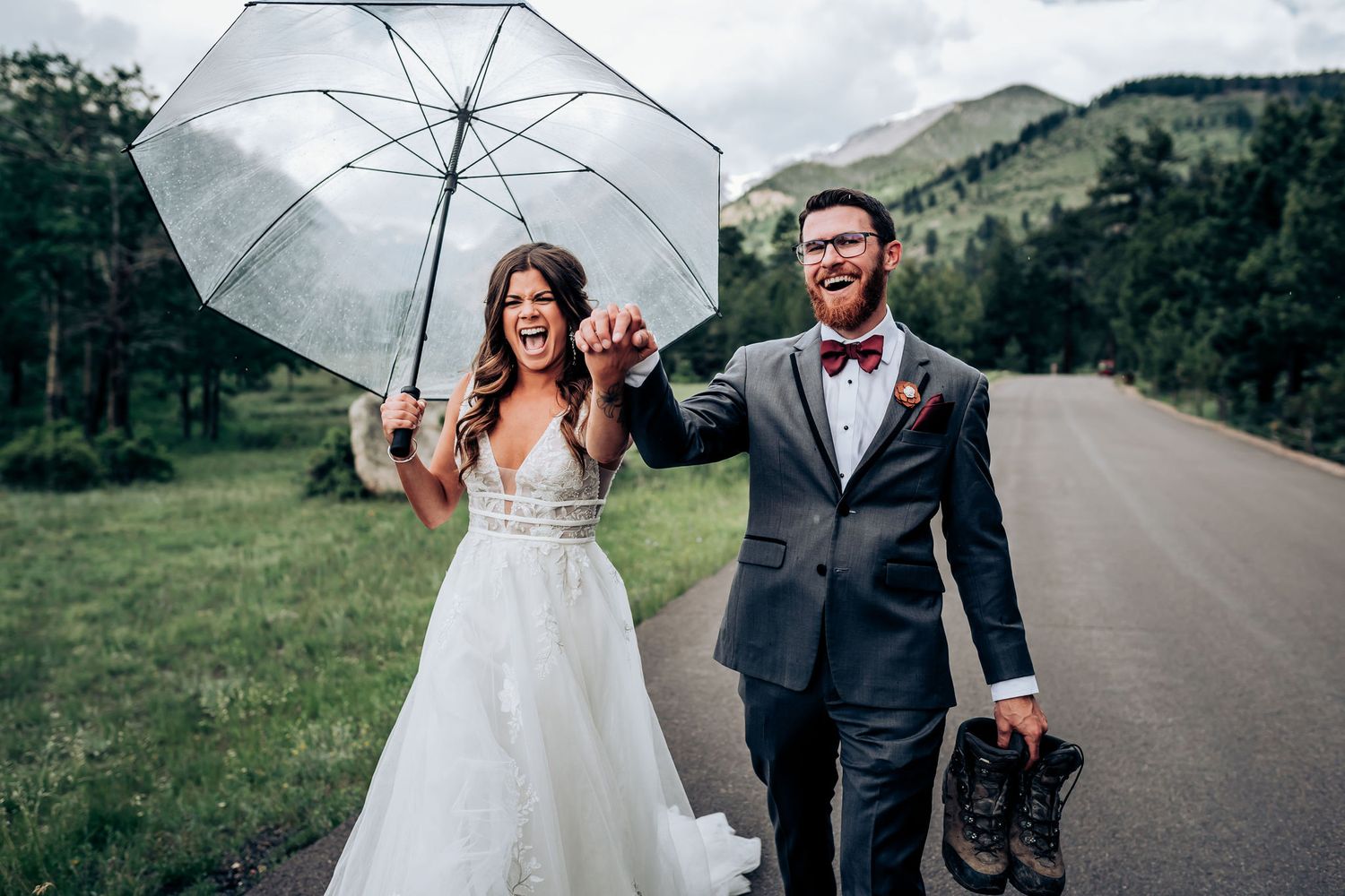A wedding couple shares a joyful moment walking down a mountain road with a white umbrella and scenic backdrop.