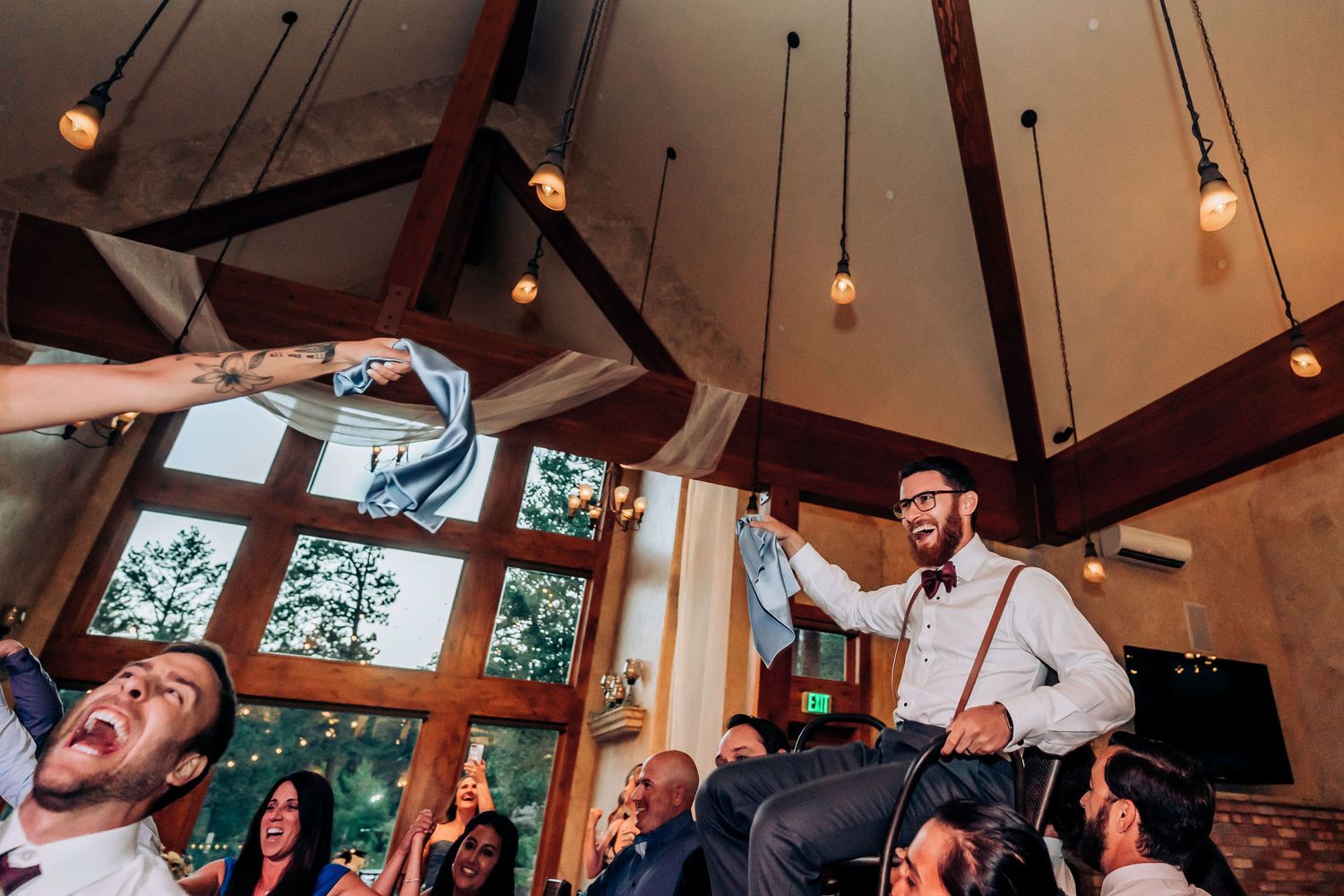 Wedding guests cheer and celebrate during a lively reception in a rustic venue with vaulted ceilings and pendant lights.