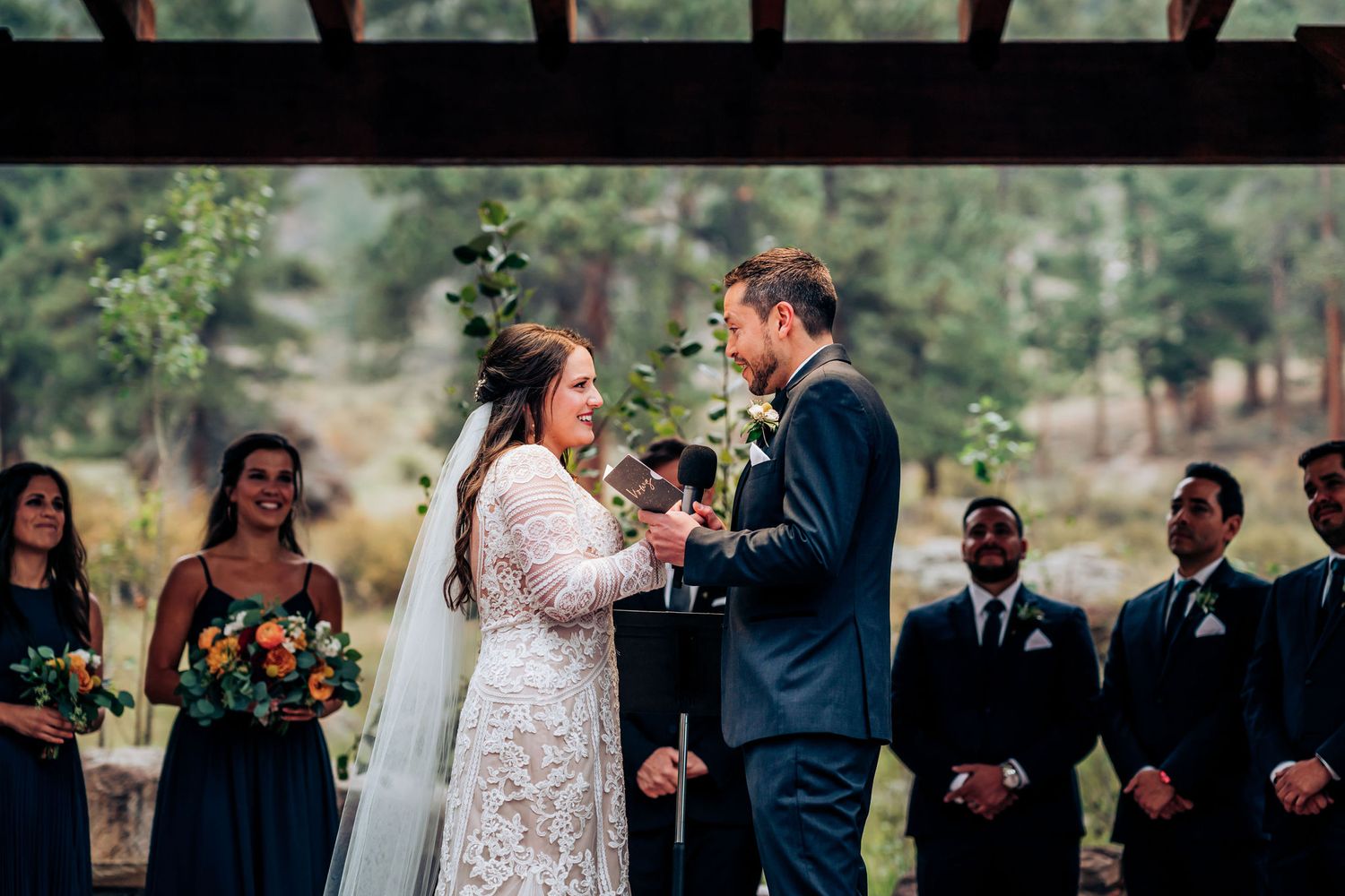 Intimate wedding ceremony moment under wooden pergola with wedding party in navy and black attire.