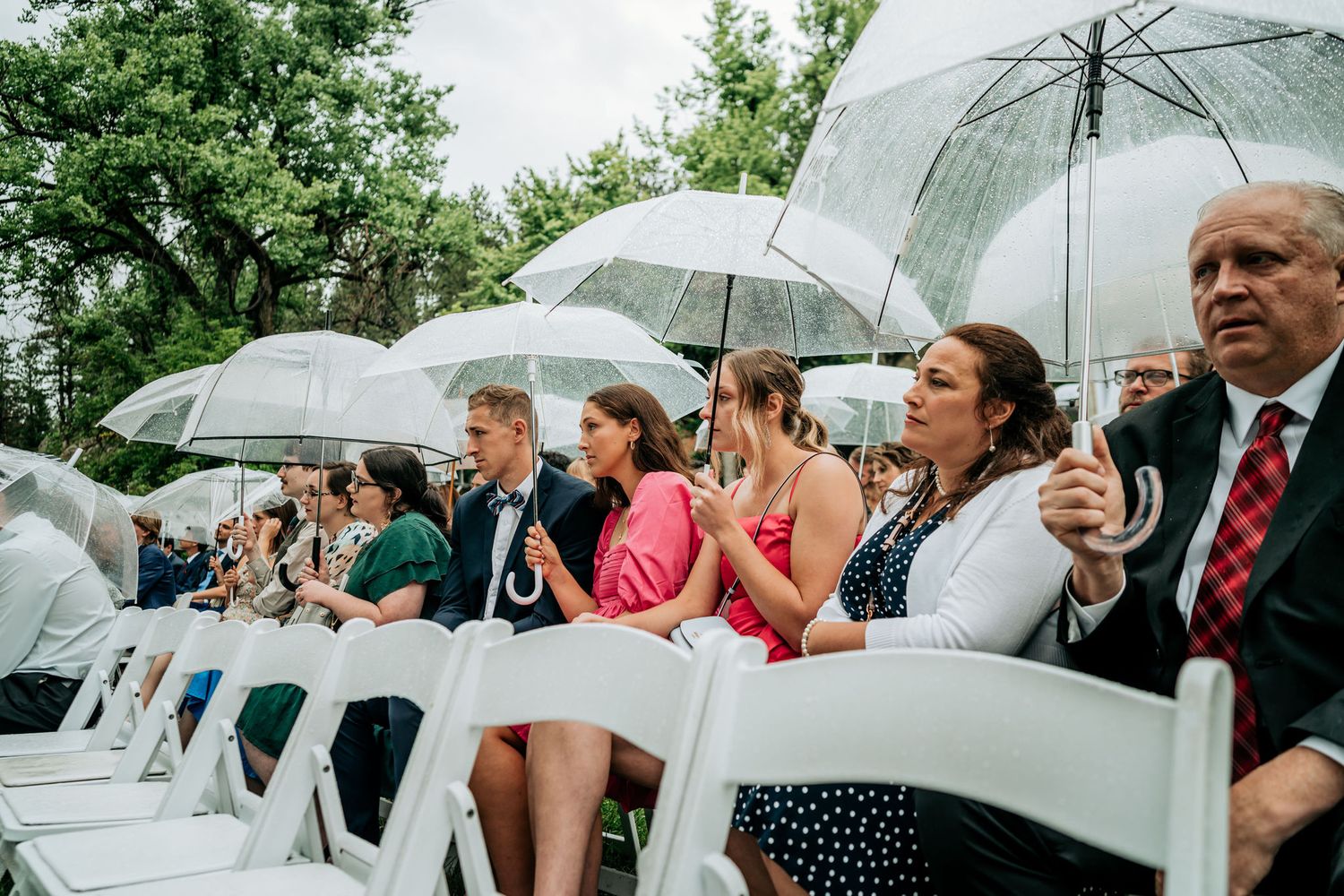 Wedding guests shelter under clear umbrellas during an outdoor ceremony on white chairs.