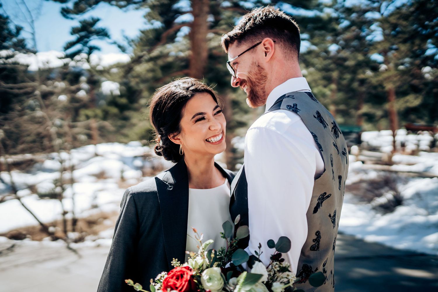 Joyful winter wedding couple sharing a candid moment among snowy pines with elegant floral bouquet.