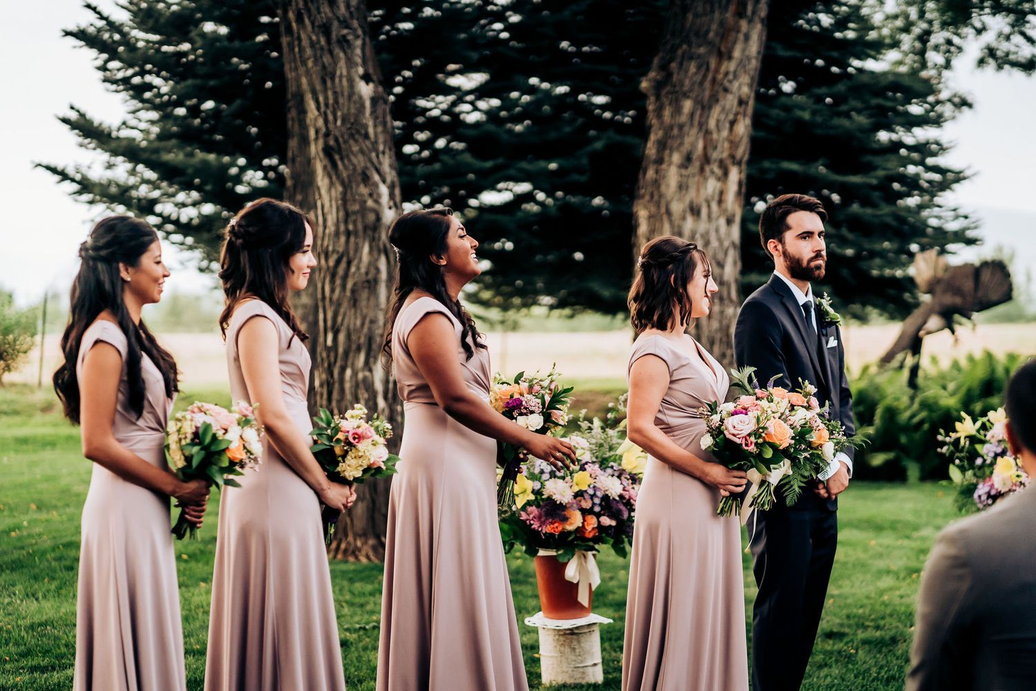 Bridesmaids in blush gowns hold bouquets during outdoor garden wedding ceremony.