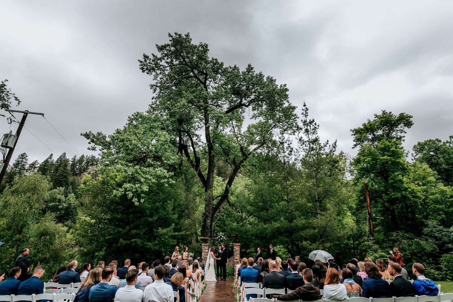 Wide shot of outdoor wedding ceremony with seated guests under cloudy skies.