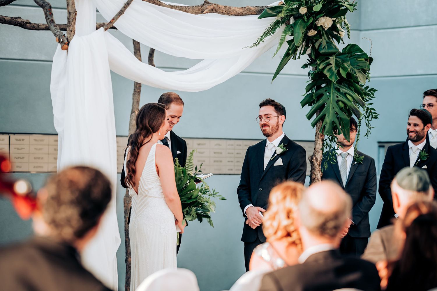 Outdoor wedding ceremony under white draped arch with lush greenery.