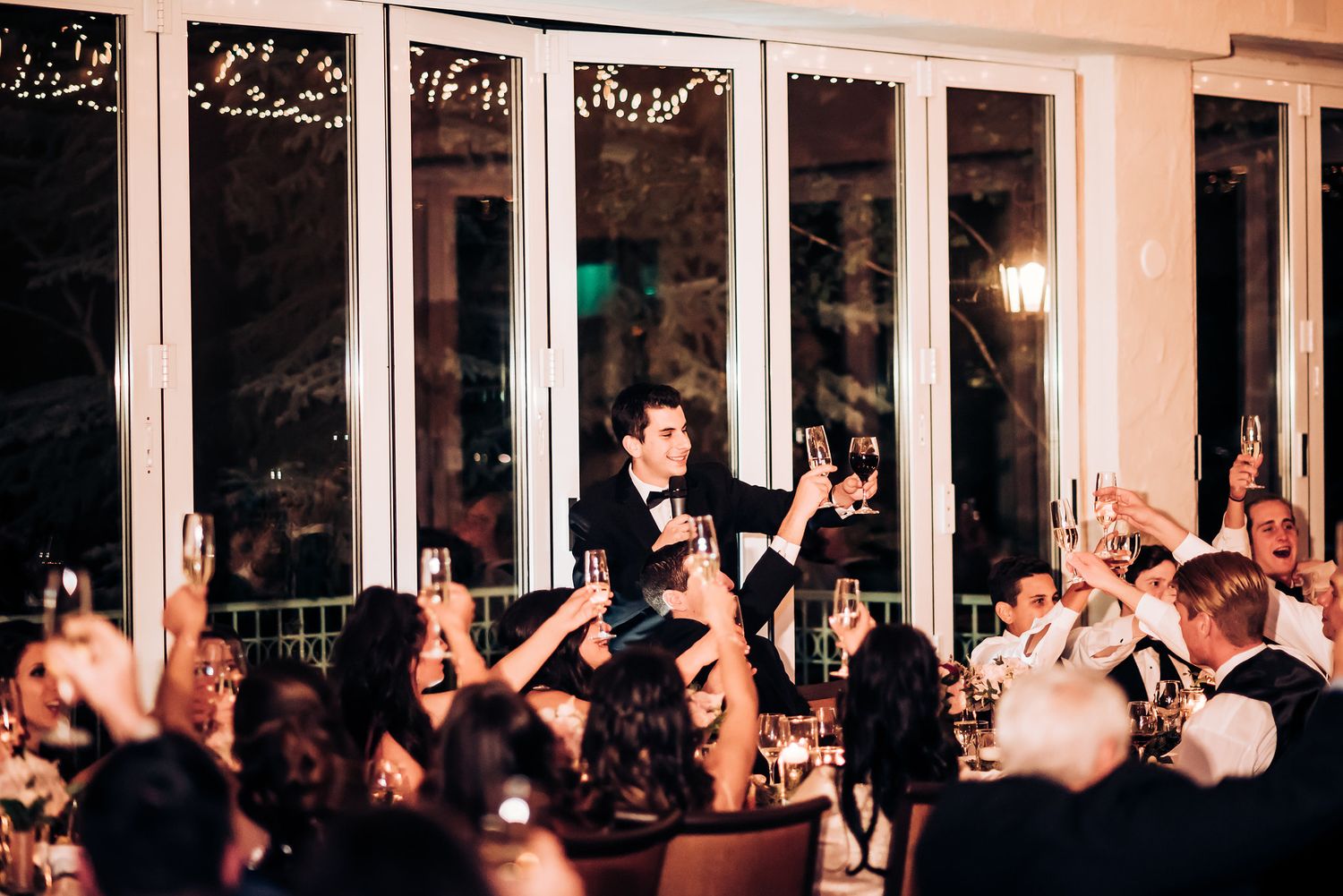 Wedding reception crowd cheers during a celebratory moment in an elegant windowed venue.