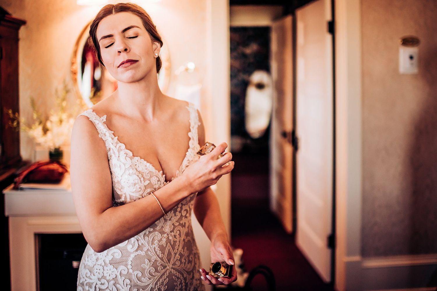 A bride in a lace wedding dress prepares for her ceremony in a warm-lit hotel room hallway.