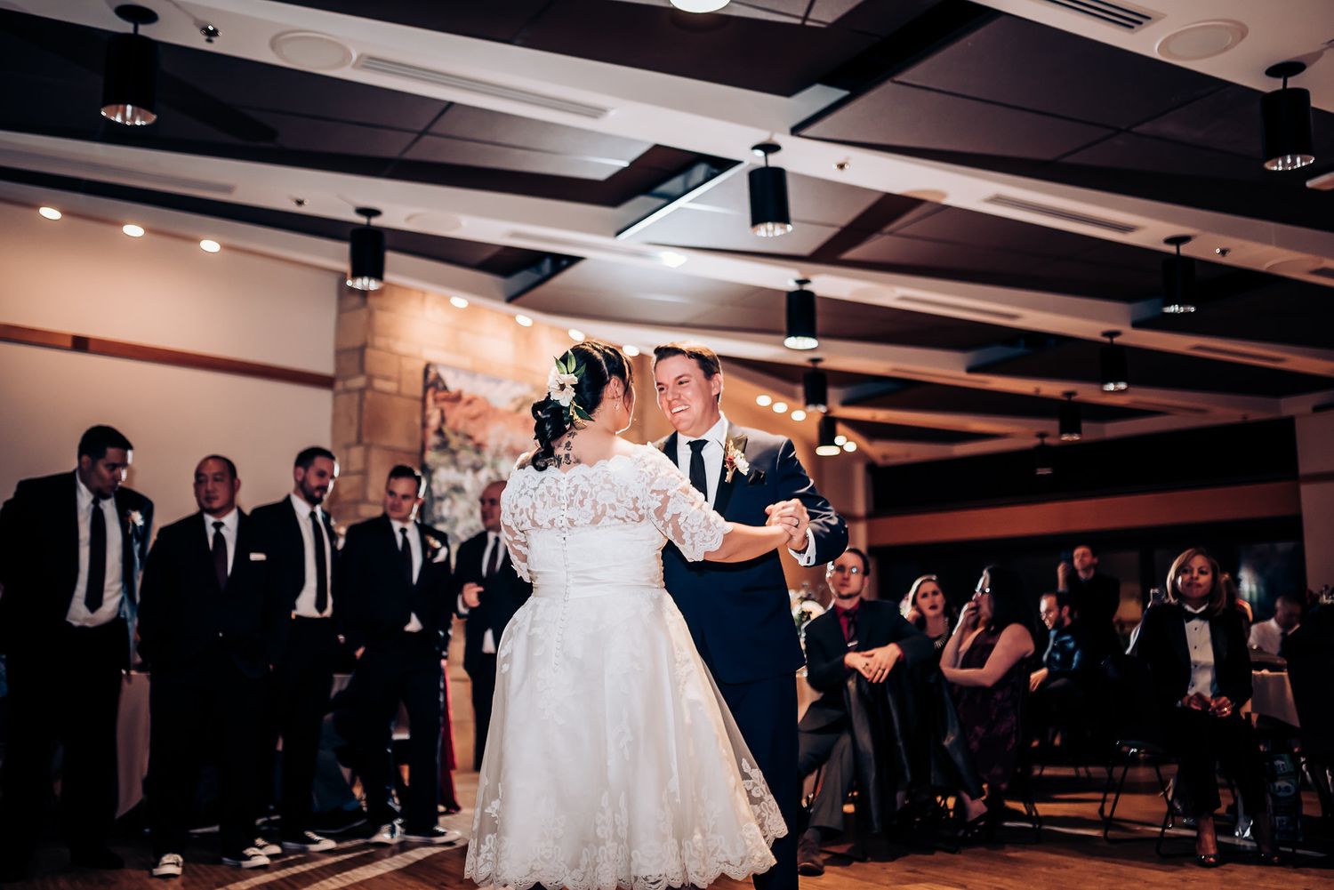 A wedding couple dances in a rustic venue with exposed beams while guests look on from the sides.