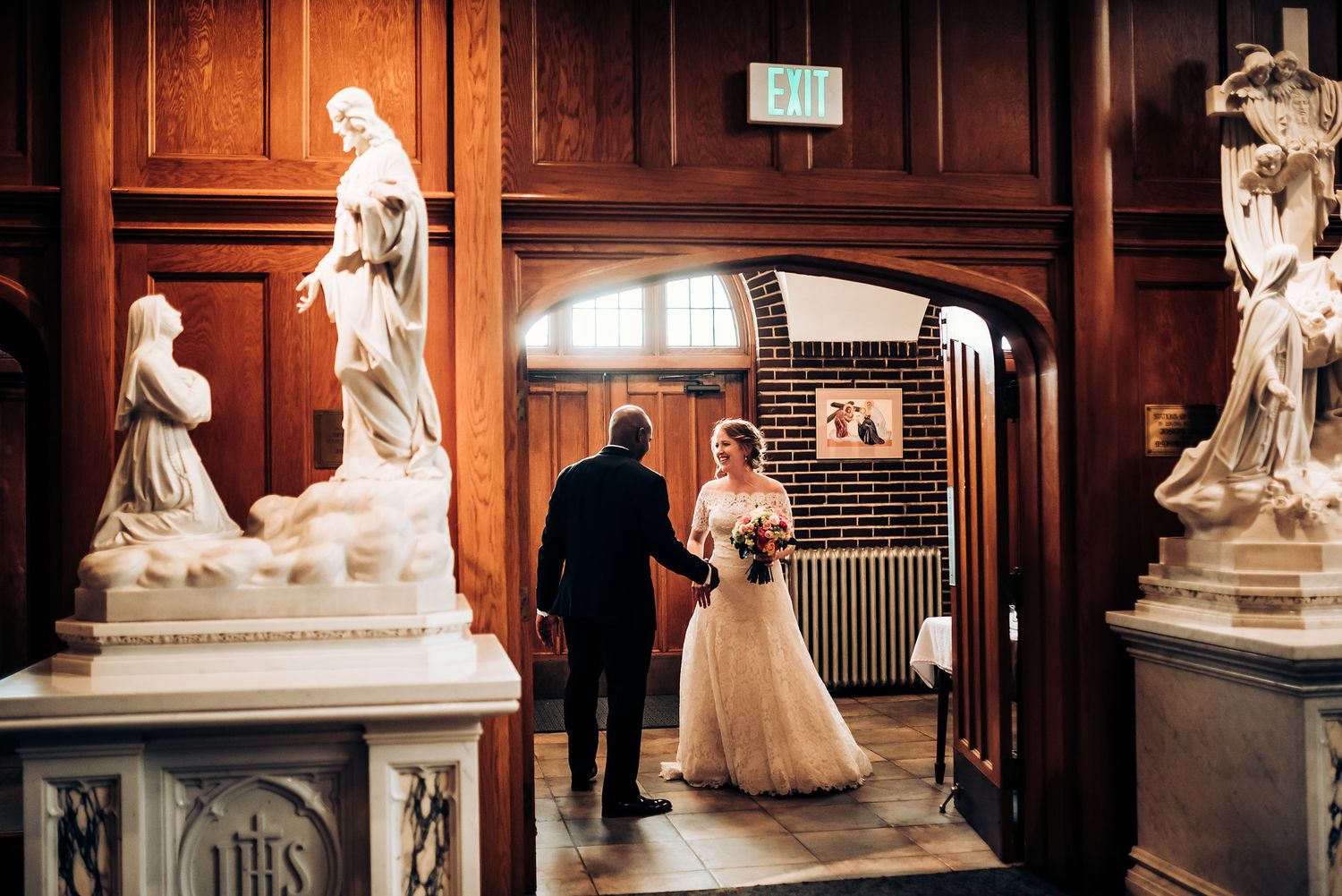 A wedding couple walks through an ornate hallway decorated with classical statues and wood paneling.