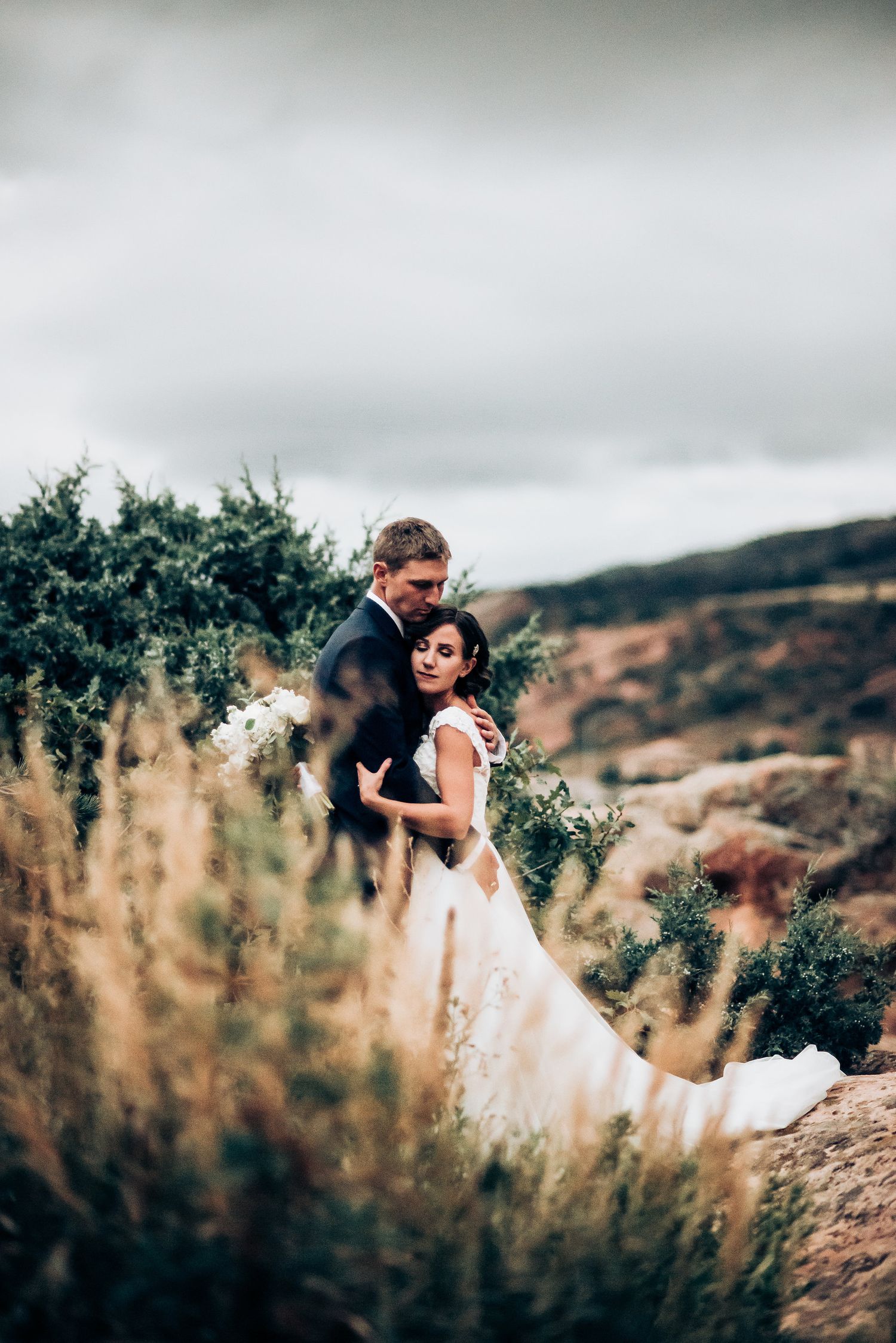 Romantic wedding portrait set against a dramatic mountain landscape with tall grass in the foreground.