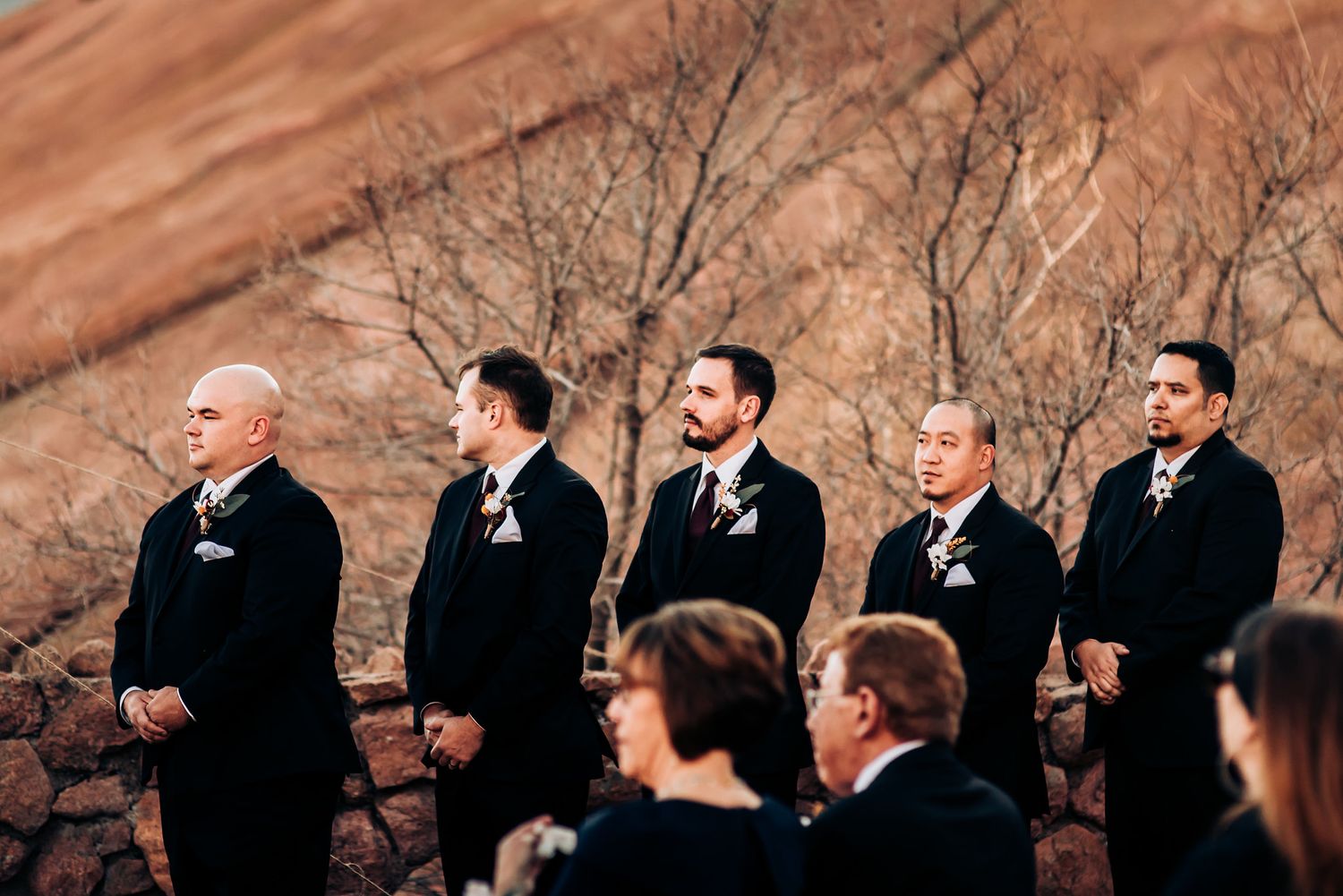 Groomsmen stand in a line against dramatic red rock cliffs during an outdoor wedding ceremony.