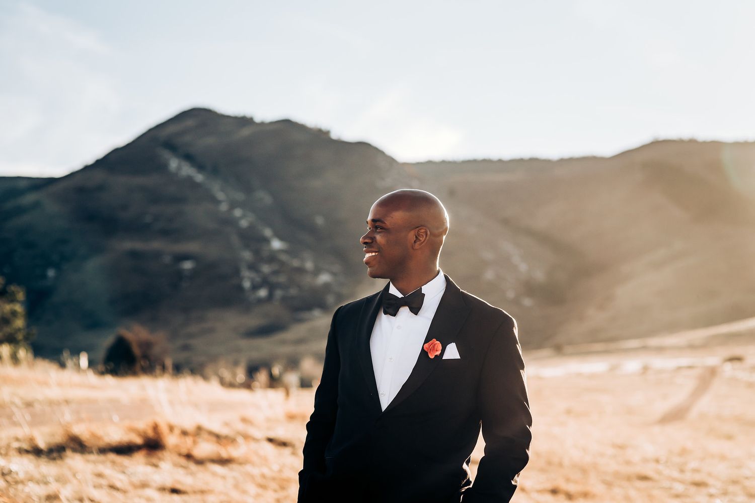 Formal groom portrait in black tuxedo and bowtie with mountains in background during golden hour.