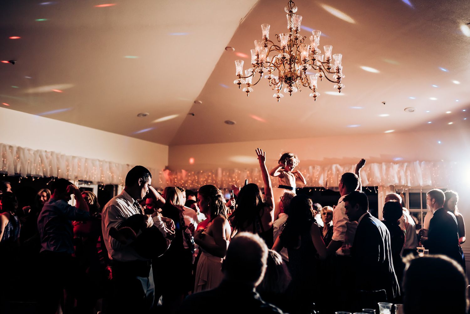 Silhouetted crowd dancing beneath a crystal chandelier at an evening celebration.