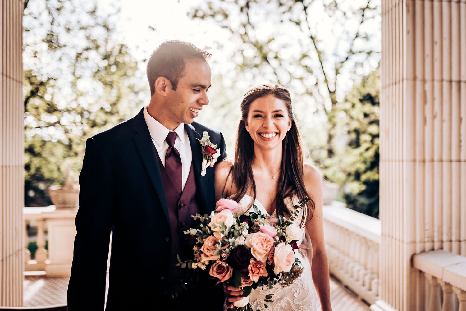 Happy couple posing on sunlit porch with pink and white floral bouquet.