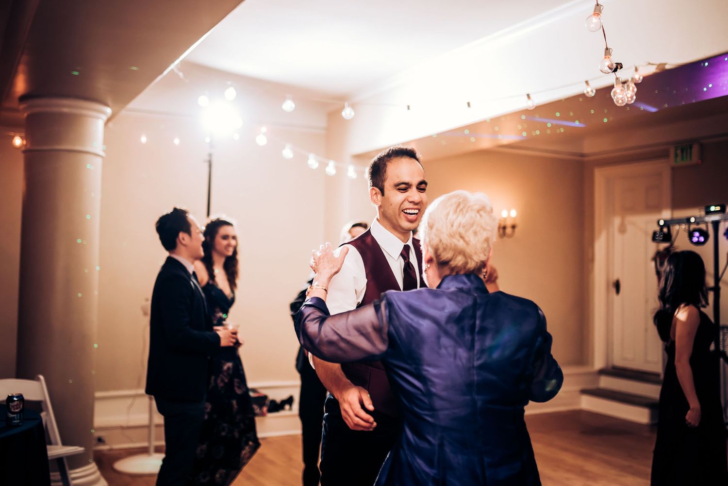Wedding guests dancing at an elegant indoor reception with string lights and columns.