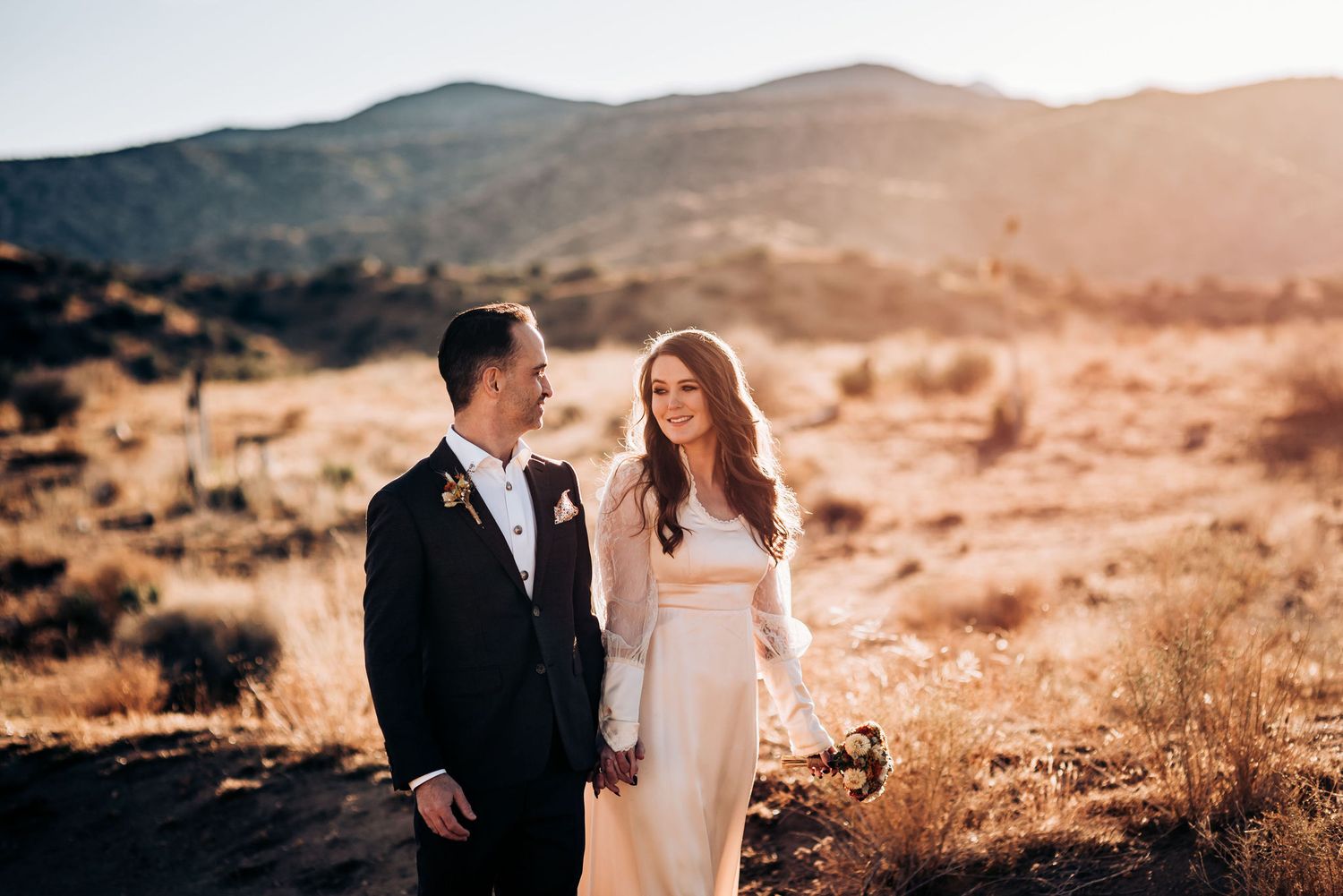 A couple in wedding attire stands together in a sun-drenched desert landscape.