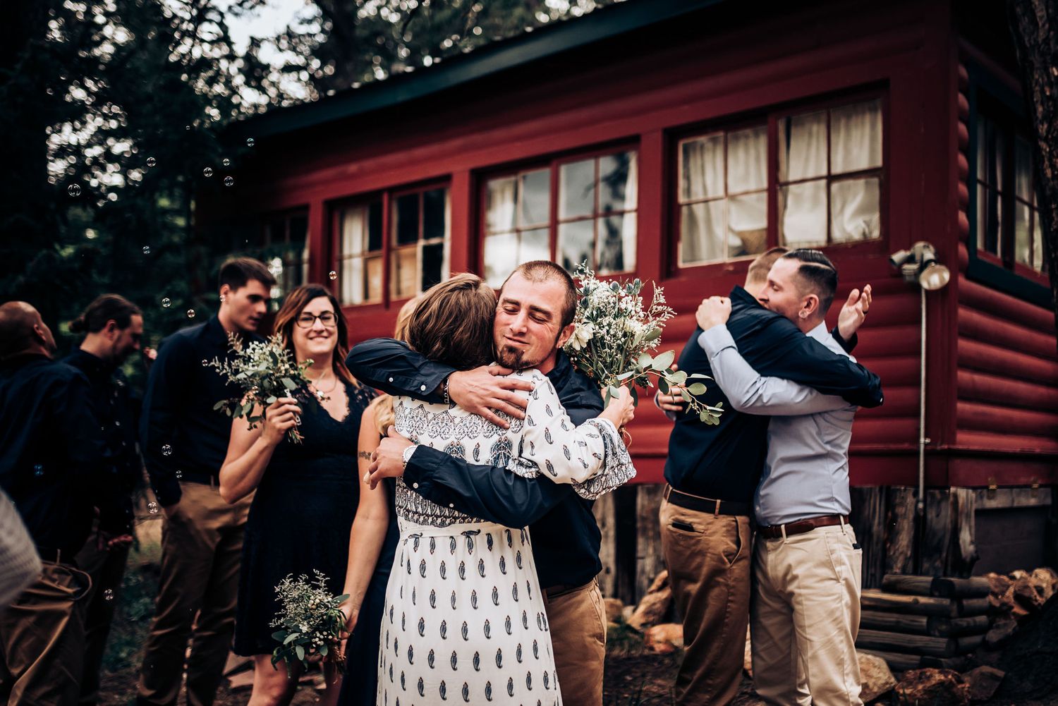 Wedding guests embrace and celebrate outside a rustic red cabin surrounded by pine trees.