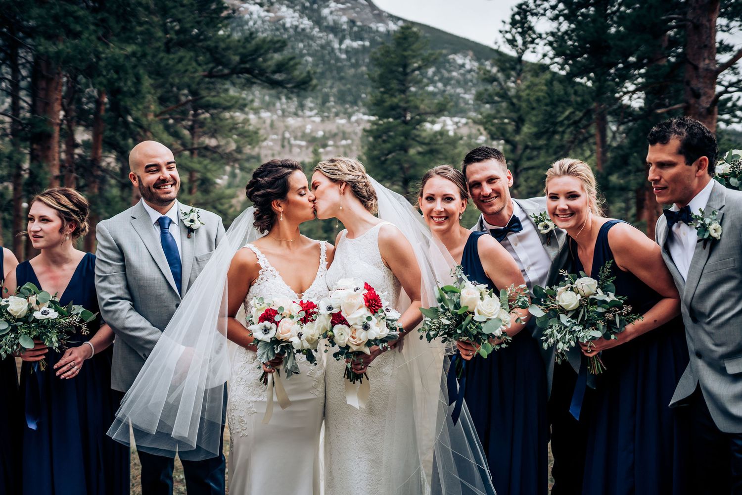 Wedding party laughs together surrounded by pine trees in a mountain setting.