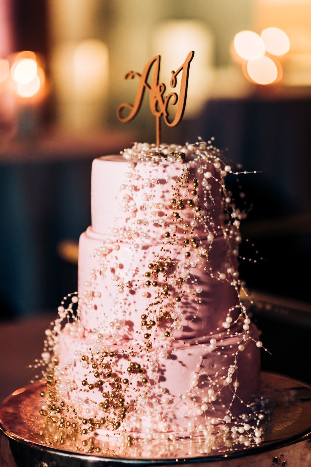Three-tiered wedding cake decorated with delicate pearls and metallic accents in soft pink lighting.