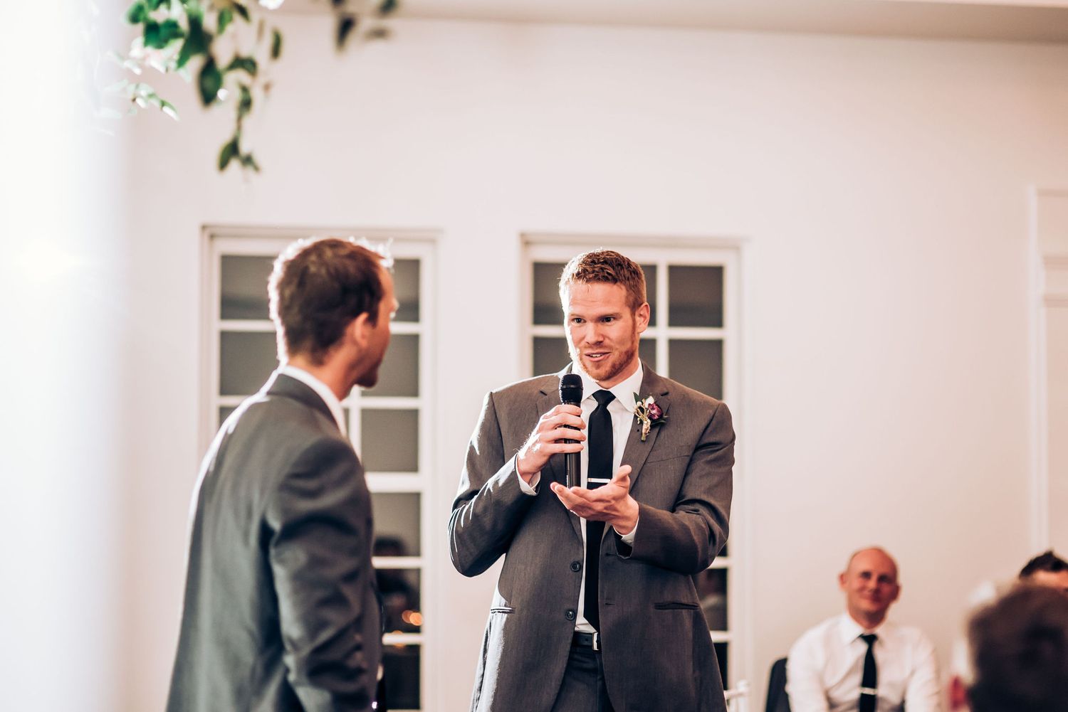 Two people in dark suits having a conversation during formal event with white paneled walls.