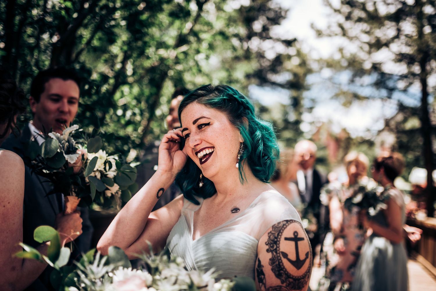 Bride with teal hair and visible anchor tattoo laughs joyfully during outdoor wedding ceremony.