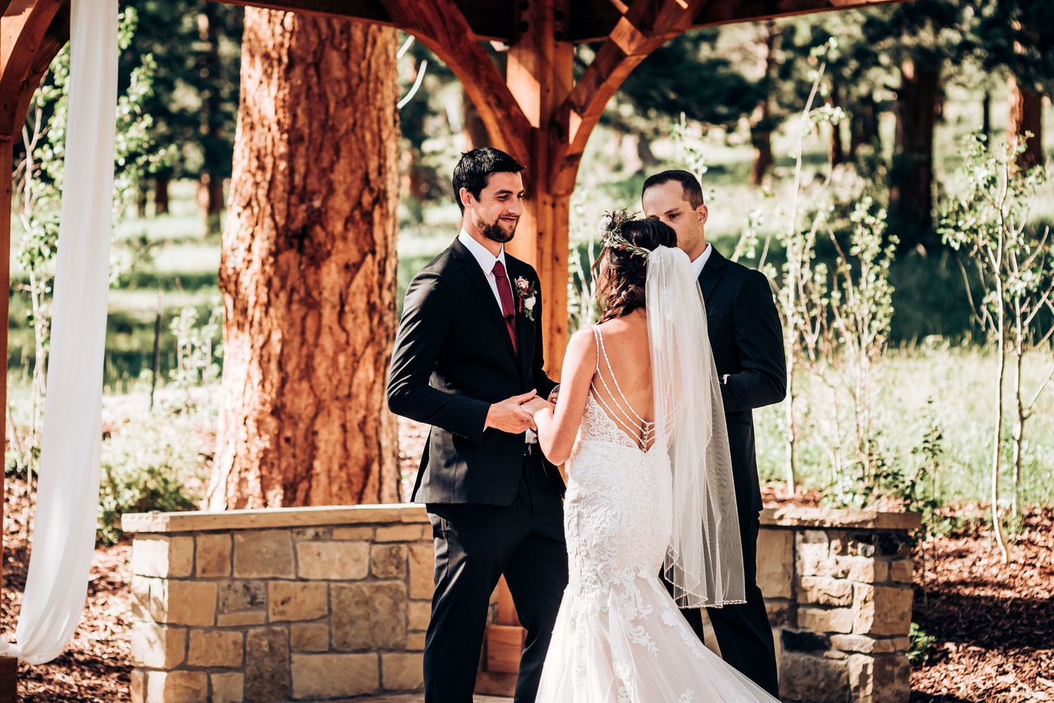 Romantic wedding ceremony in rustic outdoor setting with stone pillars and tall pine trees.
