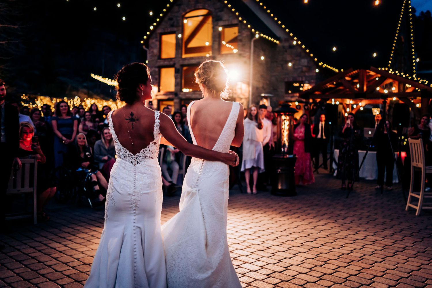 Two white wedding dresses with open backs silhouetted against evening reception lighting.