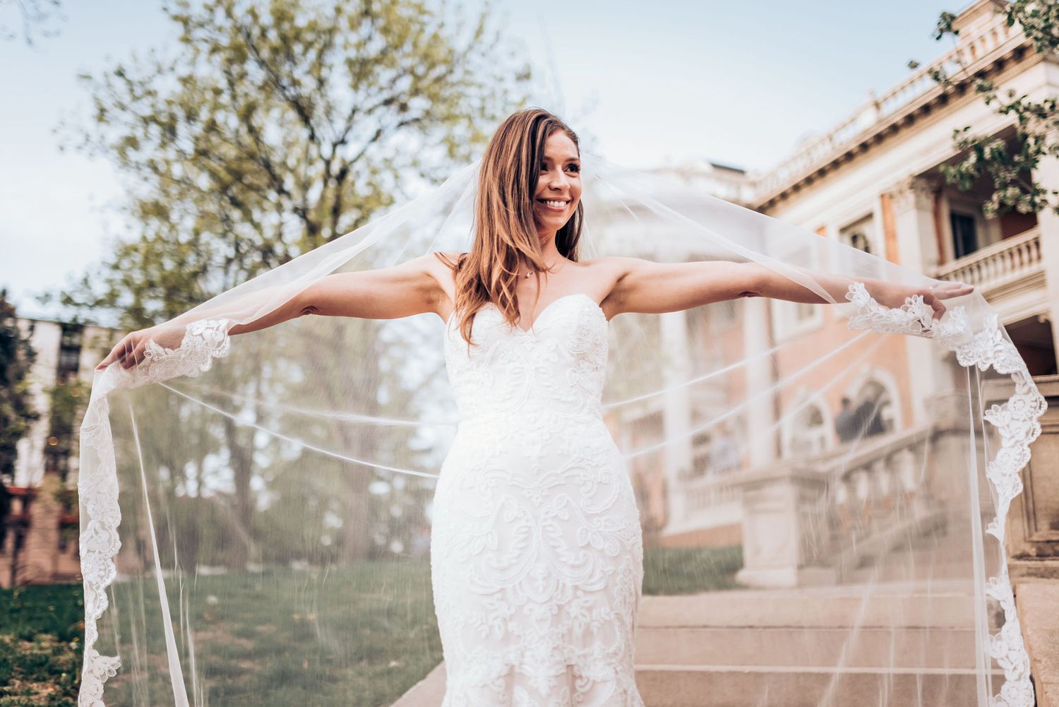 Bride in lace mermaid wedding dress twirls her long veil on stone steps of historic mansion.