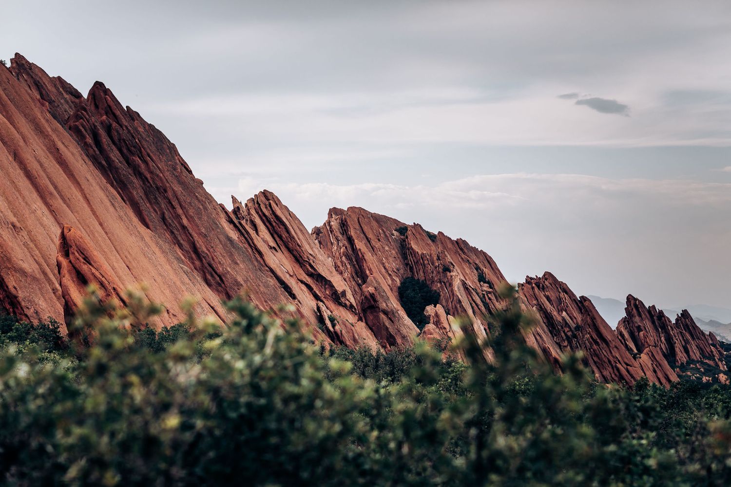 Dramatic red sandstone rock formations rise against a cloudy sky with green foliage in the foreground.