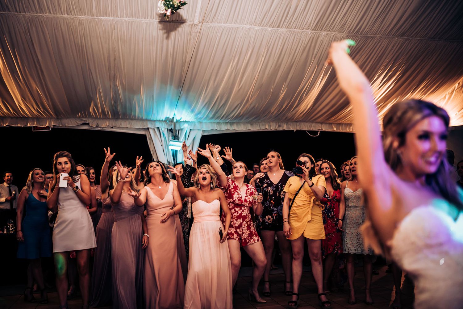 Wedding guests dance and celebrate under a draped tent ceiling with blue accent lighting at a reception party.