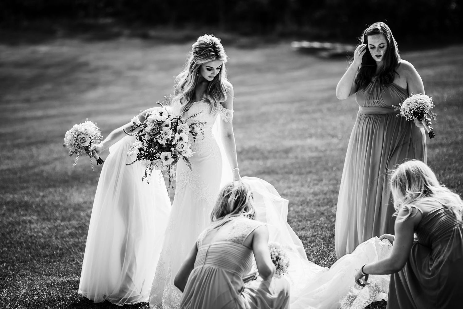 Wedding party in flowing dresses gather on a grassy field in black and white photography.