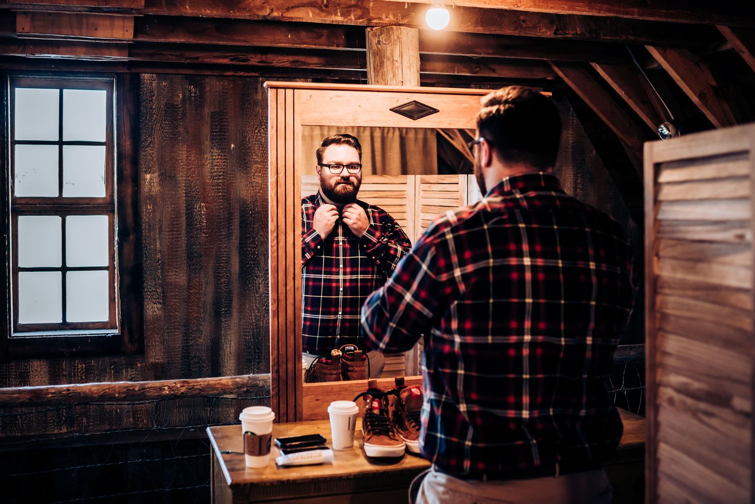 Person in plaid shirt adjusting tie while looking in rustic wooden bathroom mirror.