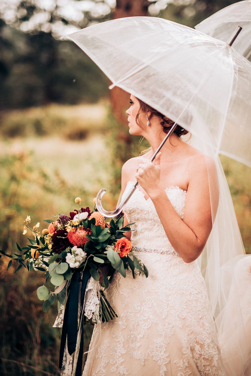 Bride holding coral bouquet stands with clear umbrella in rustic outdoor setting.