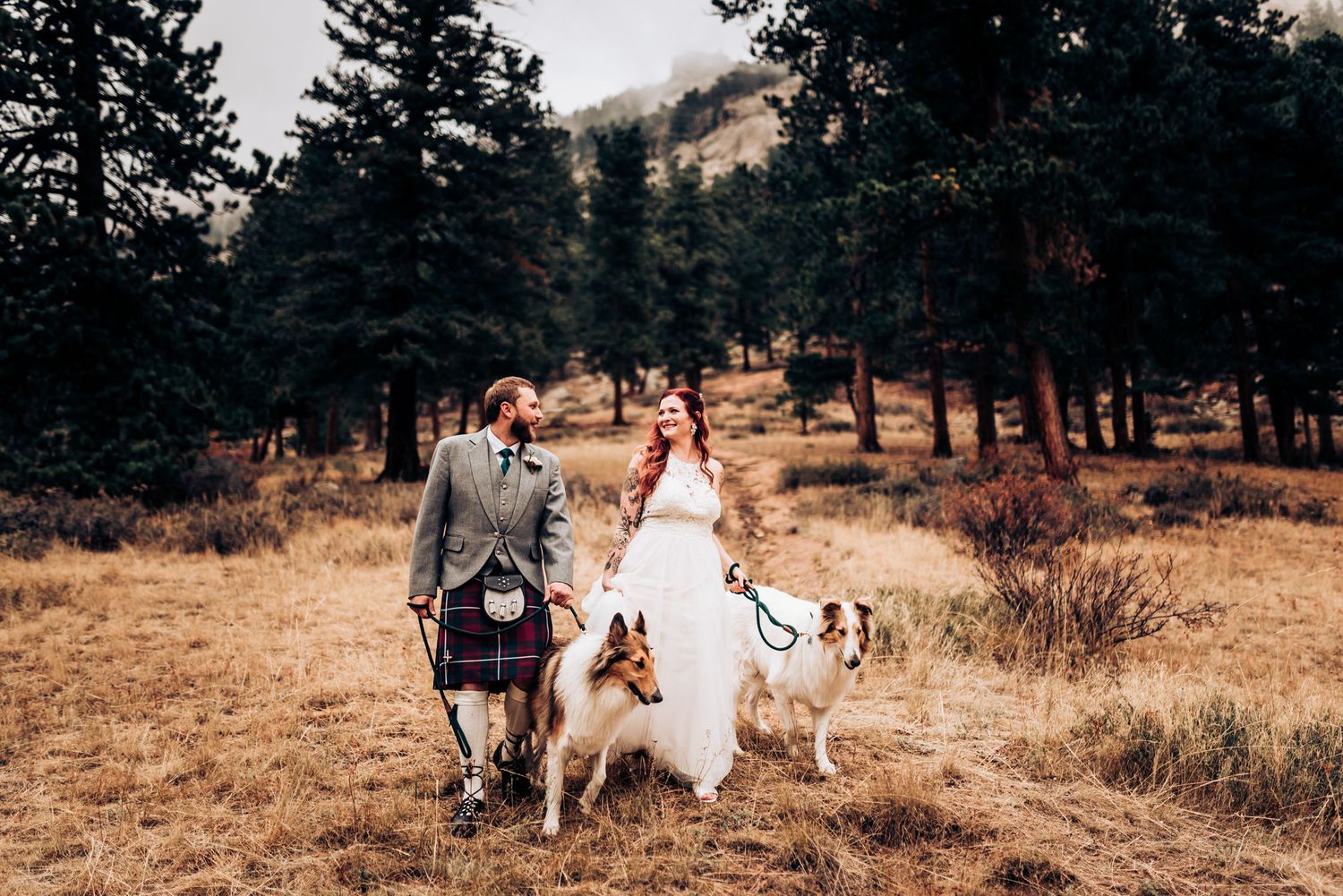 A couple walks through a pine forest meadow with their dogs on their wedding day wearing traditional Scottish attire.