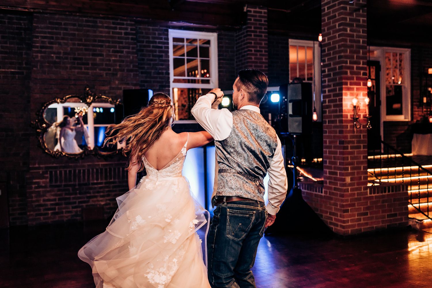 Couple dances energetically in a rustic brick-walled reception venue with warm lighting.