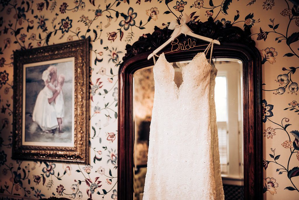 White wedding dress hanging in vintage room with floral wallpaper and ornate mirror.