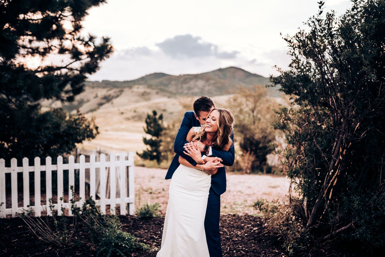 A couple shares an intimate moment in their wedding attire against a mountain backdrop with white picket fence.